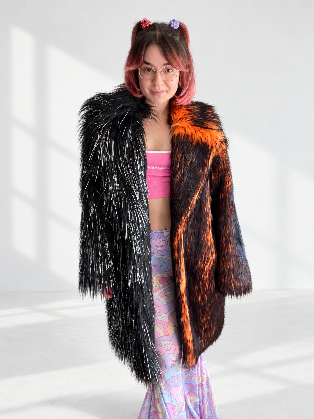 A model wearing a long, oversized faux fur coat with a split-color design: one side in black shiny tinsel fur and the other in fiery orange and black shaggy fur, photographed from the front.