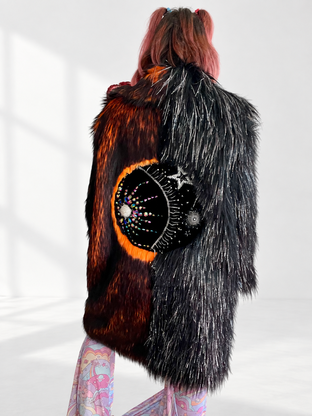 Back view of a long, oversized faux fur coat with fiery orange and black shag fur on one side and shiny black tinsel fur on the other, featuring a large hand-sewn rhinestone sun and moon design.