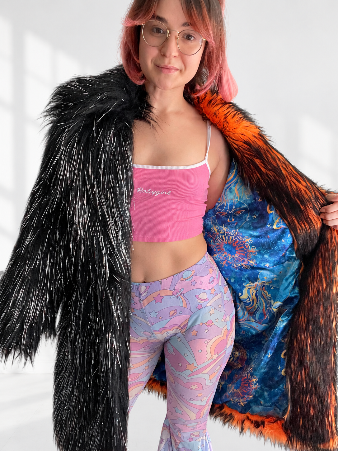 A model holding open a black and fiery orange shaggy faux fur coat, revealing its vibrant blue celestial satin lining with colorful sun, moon, and star designs.