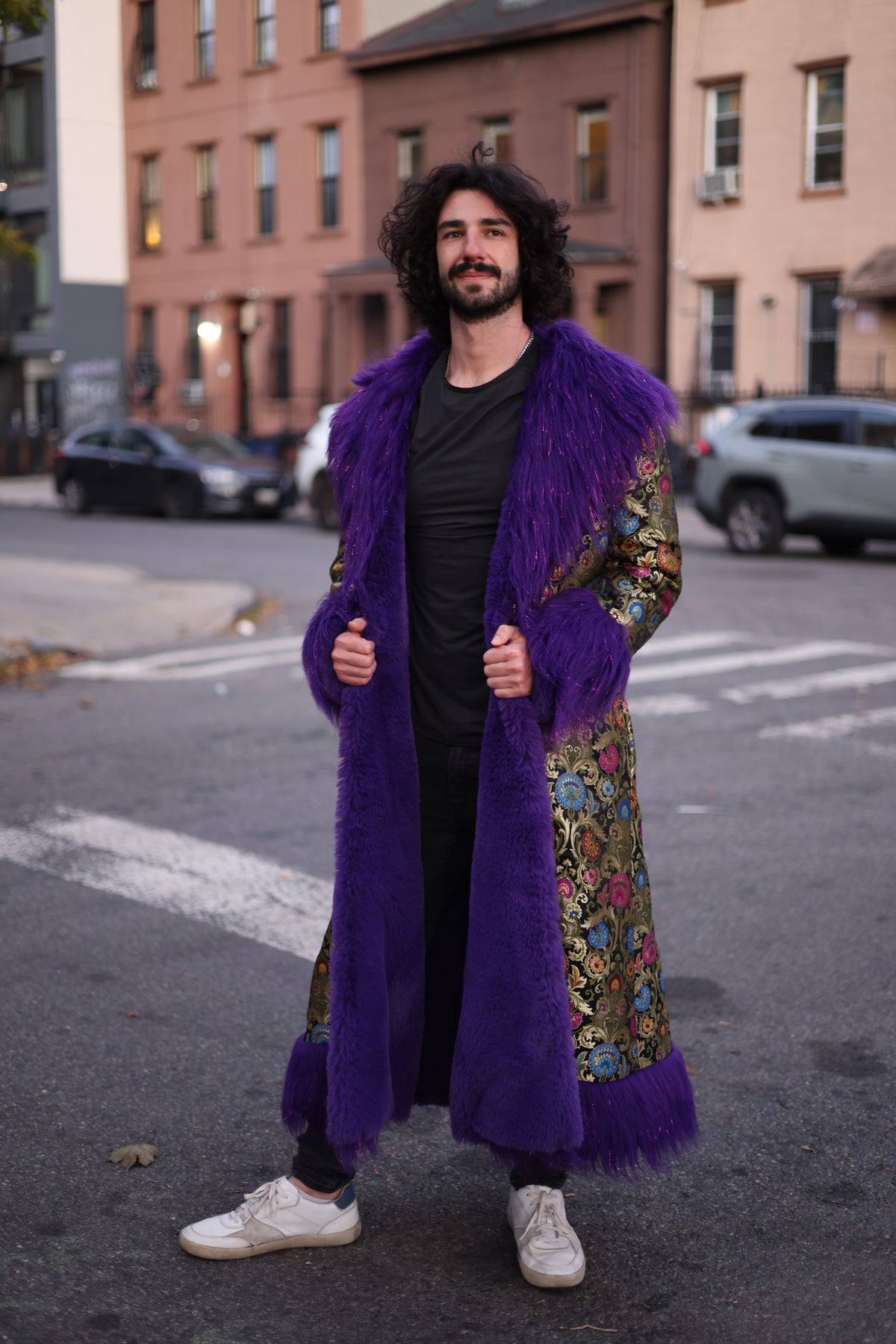 Front view of a male model wearing the purple full-length silk brocade coat with colorful floral patterns, oversized purple faux fur collar, cuffs, and front trim on a city street.