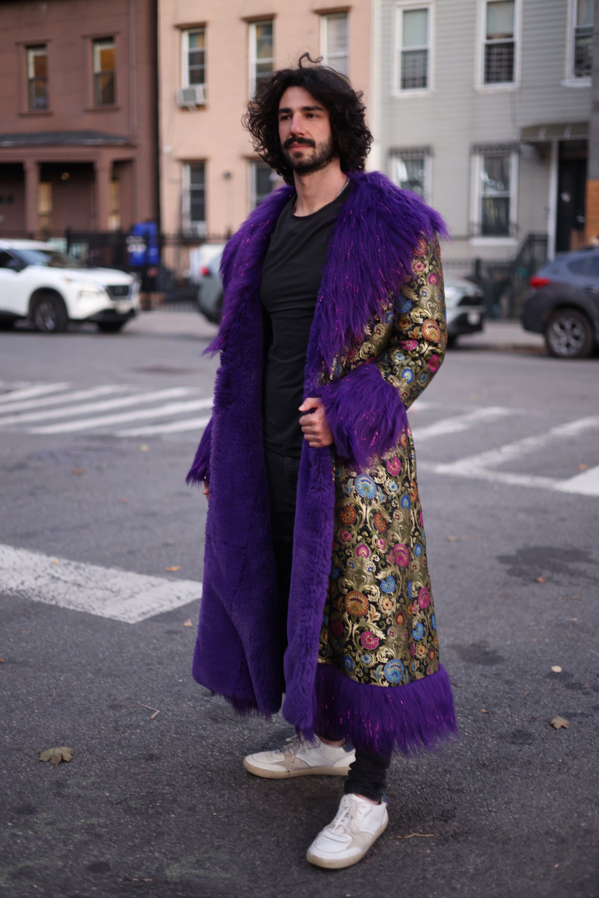 Male model wearing the PURPLE RAIN full-length silk brocade coat with colorful floral patterns, oversized purple faux fur collar, cuffs, and front trim, shown from a side angle on a city street.