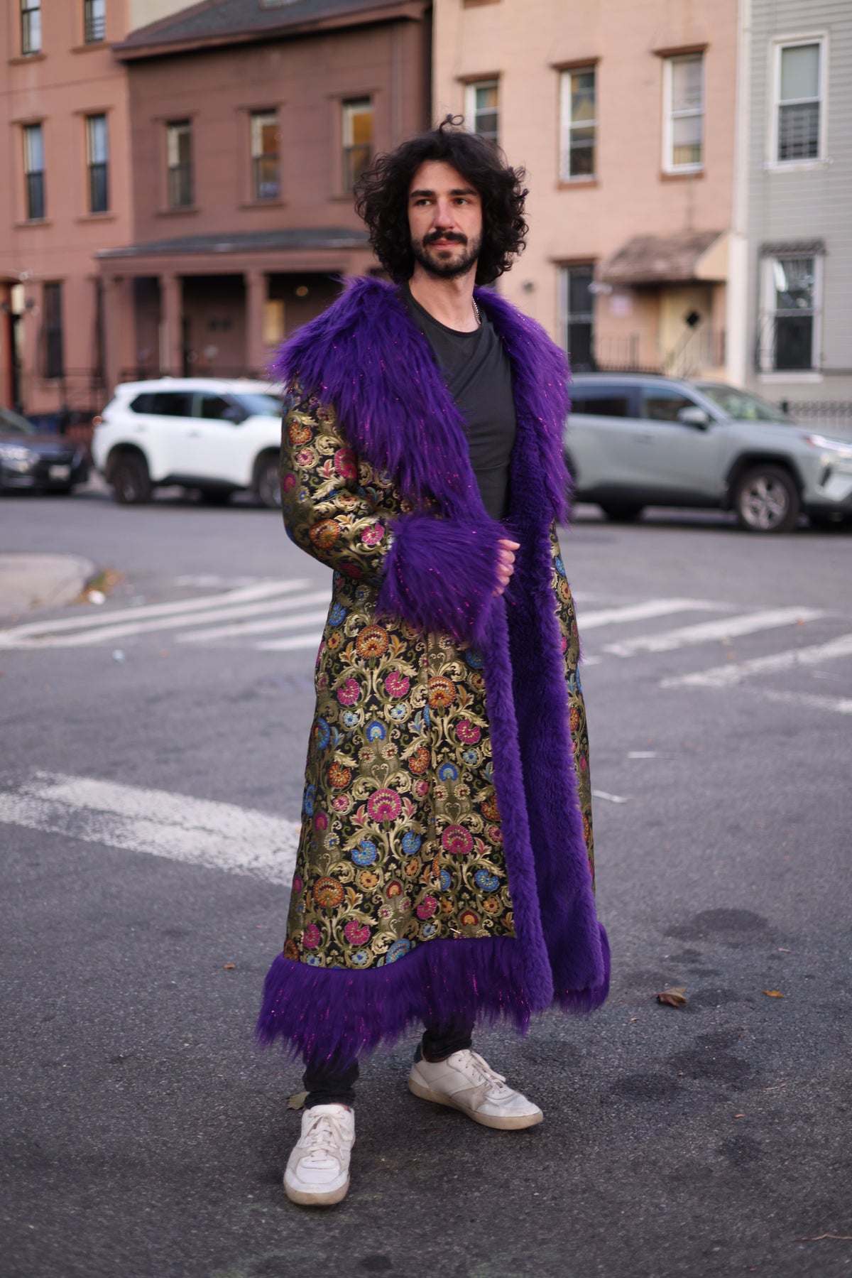 Male model wearing the PURPLE RAIN full-length silk brocade coat with a colorful floral pattern, oversized purple faux fur collar, cuffs, and front trim on a city street.
