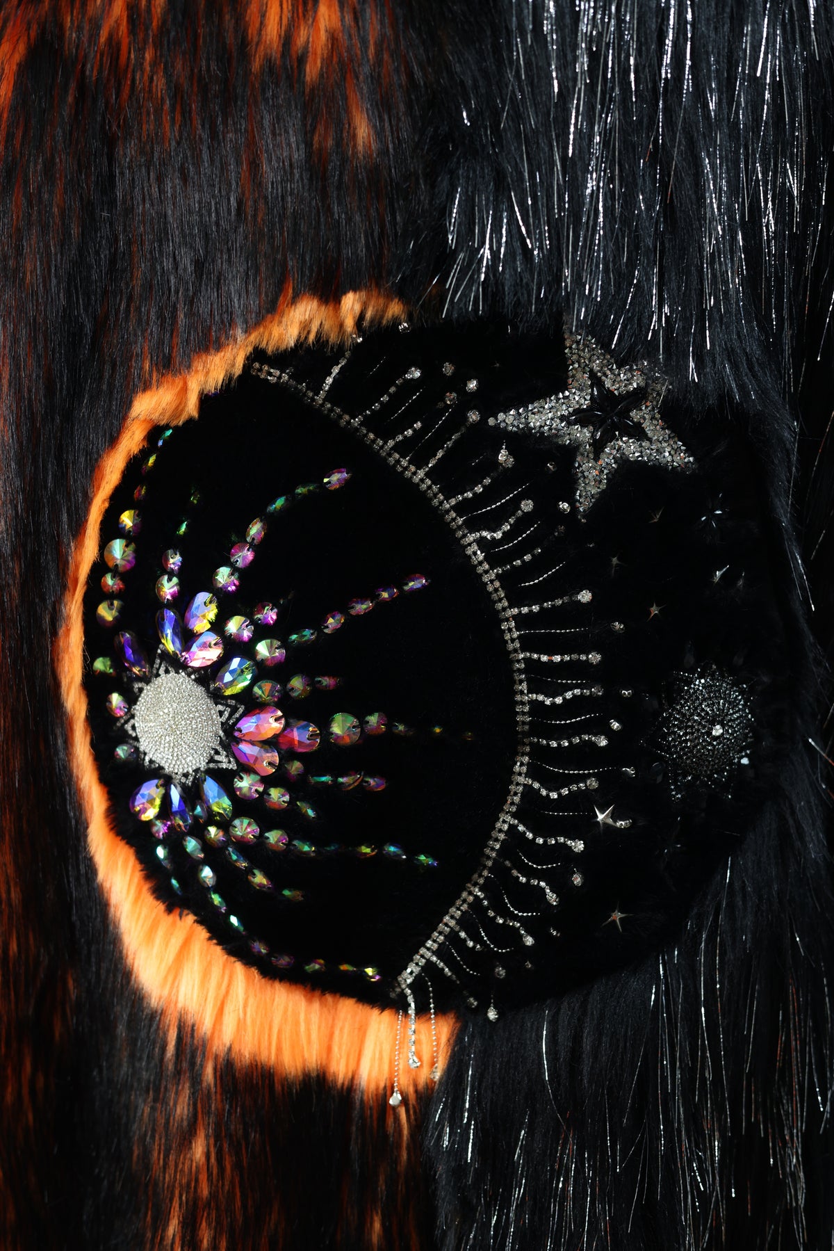 Close-up of the ECLIPSE faux fur coat's hand-sewn rhinestone sun and moon design, featuring iridescent crystals, silver stars, and contrasting black and fiery orange fur.